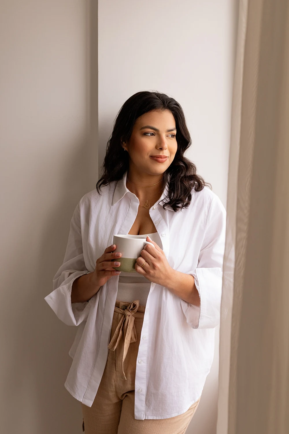 Fernanda Coutinho segurando uma xícara de café junto à janela.